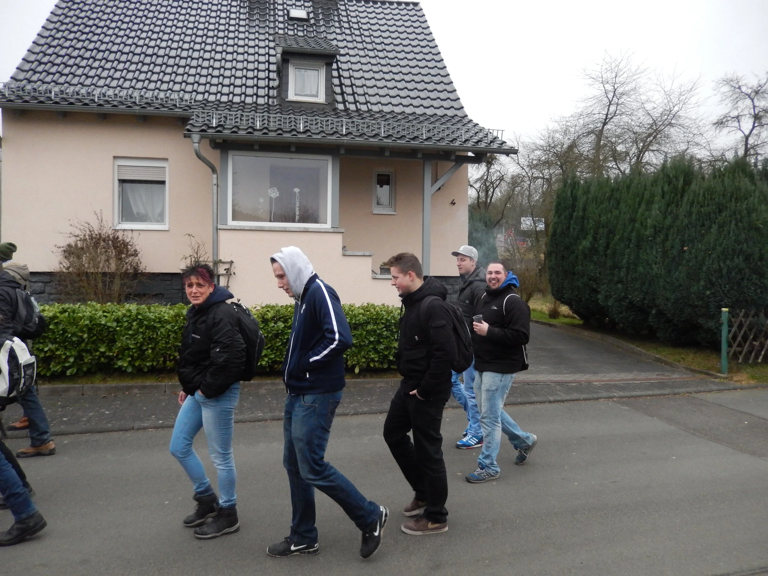 Eine Gruppe junger Menschen geht fröhlich die Straße entlang, im Hintergrund ein Haus mit einem Giebeldach und Bäumen.