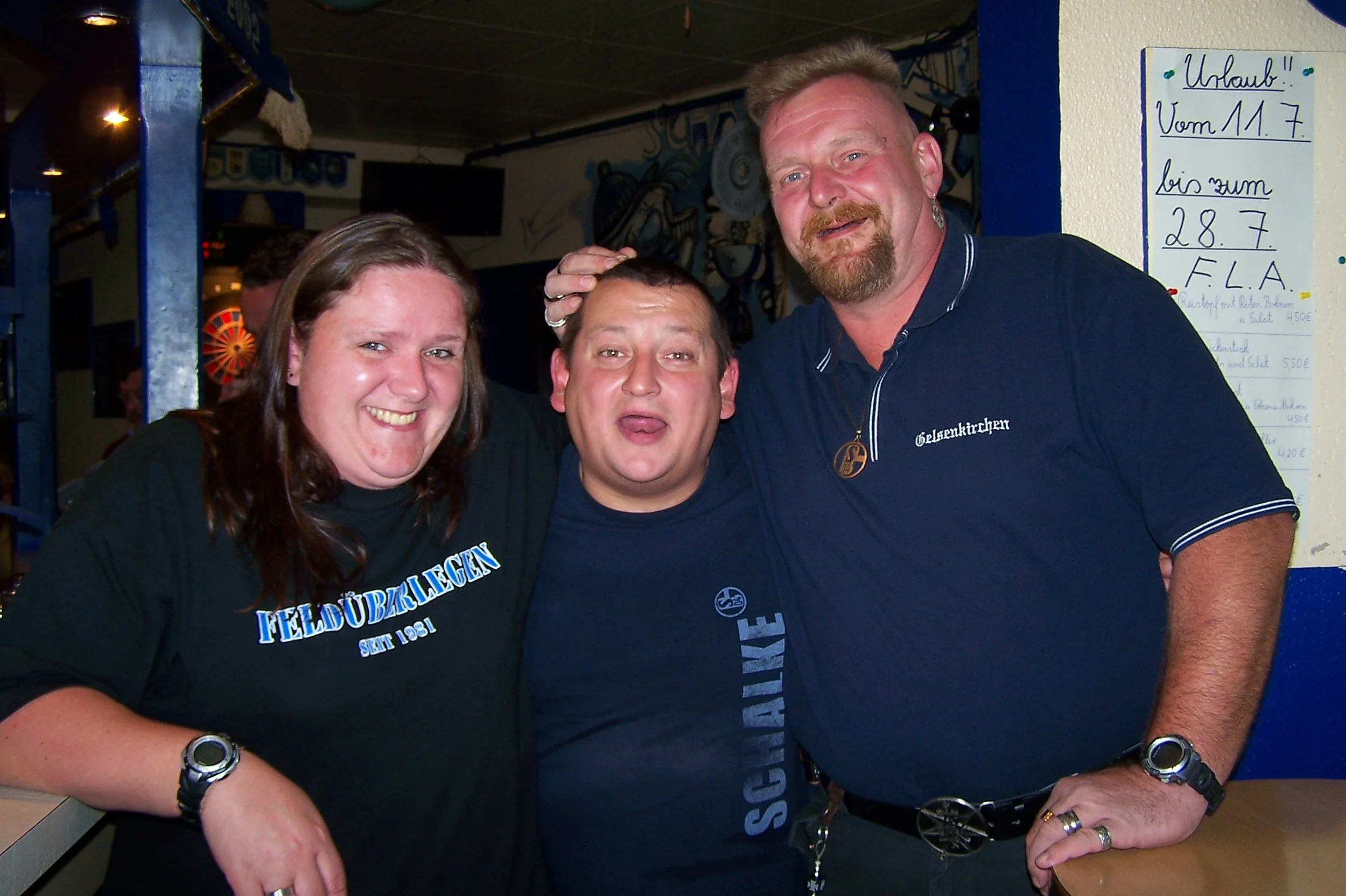 Drei Menschen, die glücklich lachen und sich umarmen, in einer Bar oder einem Club, mit einer schwarzen Wand im Hintergrund und einem weißen Tafel mit handgeschriebenen deutschen Texten an der Wand.