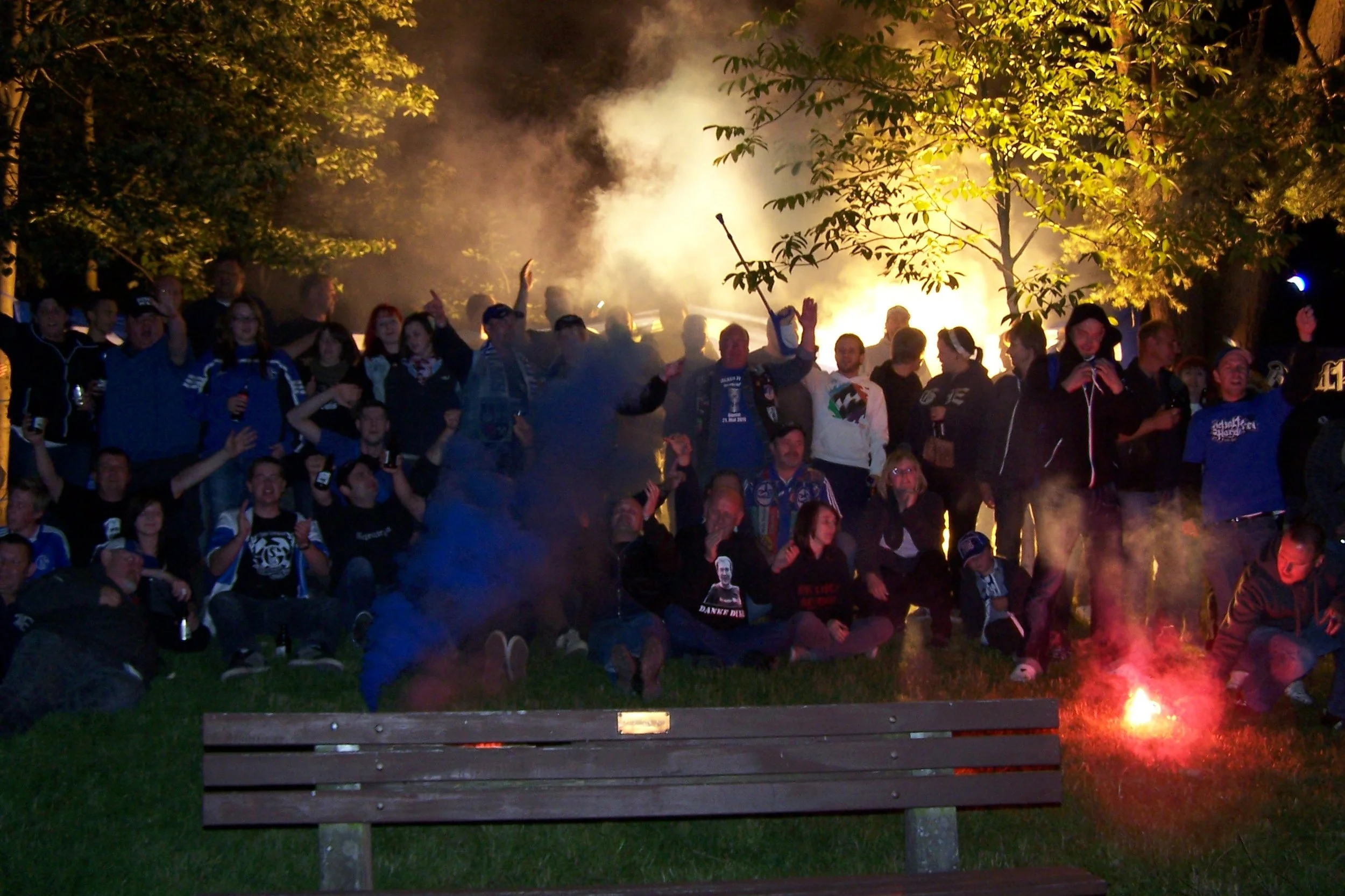 Gruppe von Menschen, die nachts um ein Lagerfeuer versammelt sind, mit Rauch und Flammen, umgeben von Bäumen und im Dunkeln, einige Leute lachen und halten Getränke in der Hand.
