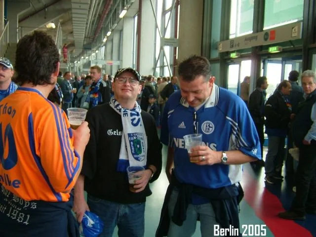 Fußballfans in einer Halle, einige tragen Trikots und scarves, im Vordergrund lachen zwei Männer mit Getränken. Text: Berlin 2005.