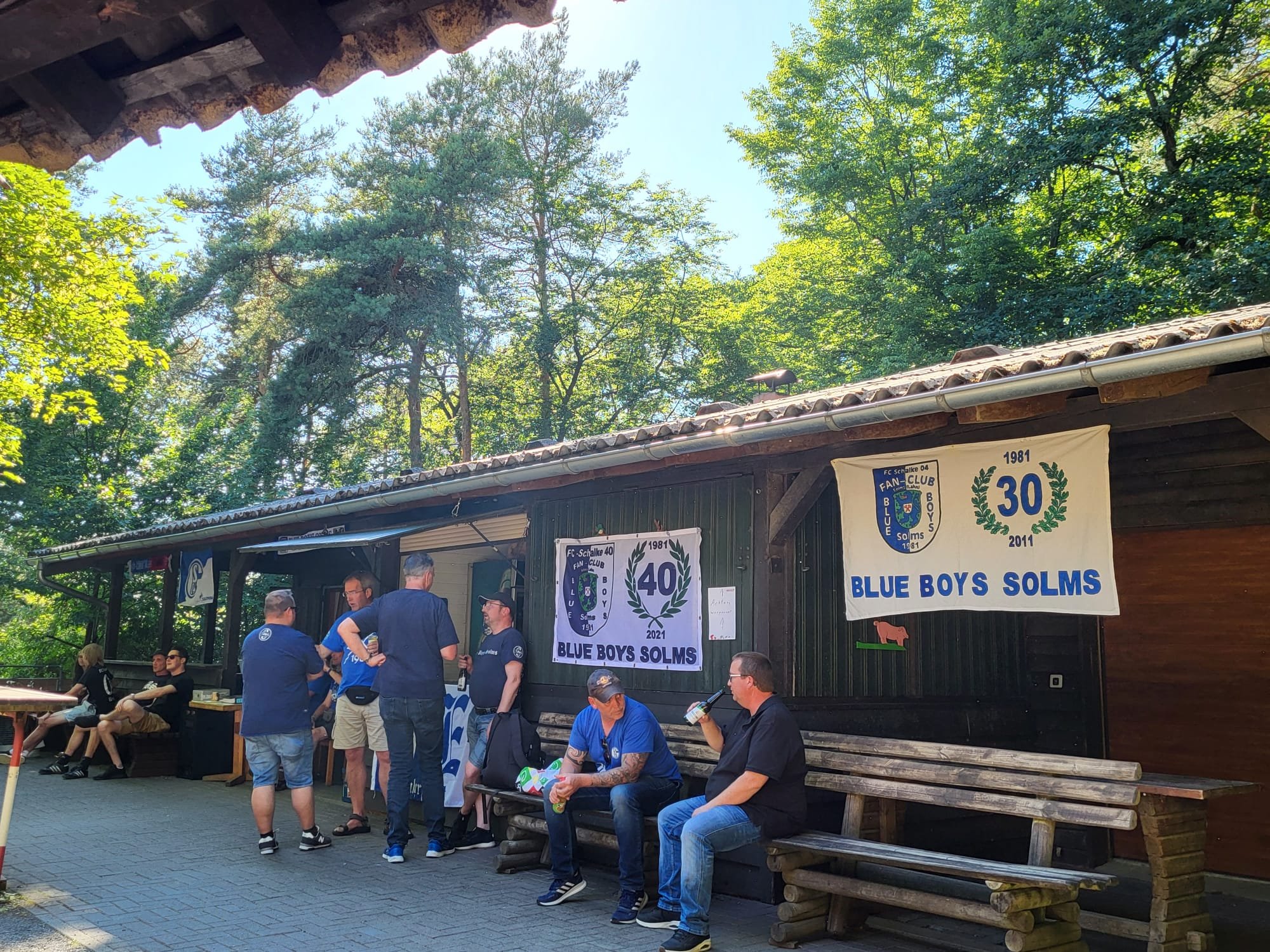 Gruppe von Menschen vor einem Holzgebäude mit Banner, das die Jahre 1981, 2011 und 2021 zeigt, mit der Aufschrift 'BLUE BOYS SOLMS', im Freien unter Bäumen an einem sonnigen Tag.