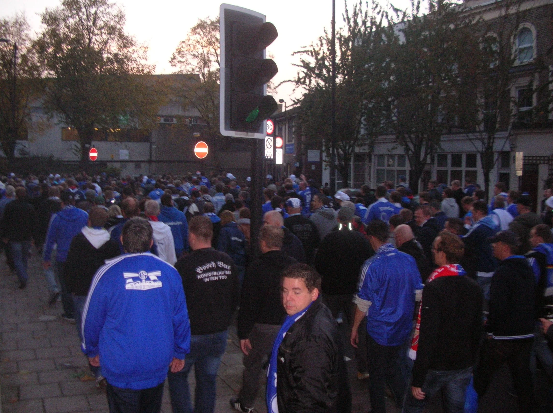 Straßenkreuzung mit Stopplicht und Menschenmenge, die in einer Stadt versammelt sind, mit Bäumen und alten Gebäuden im Hintergrund.