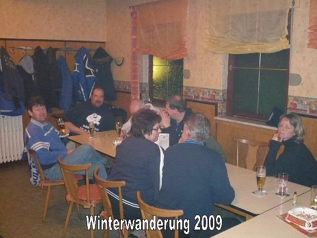 Menschen sitzen an einem langen Tisch in einem rustikalen Raum, vermutlich bei einer Winterwanderung im Jahr 2009, mit Getränken und einem Brettspiel. Im Hintergrund sind Mäntel an einer Garderobe sichtbar.
