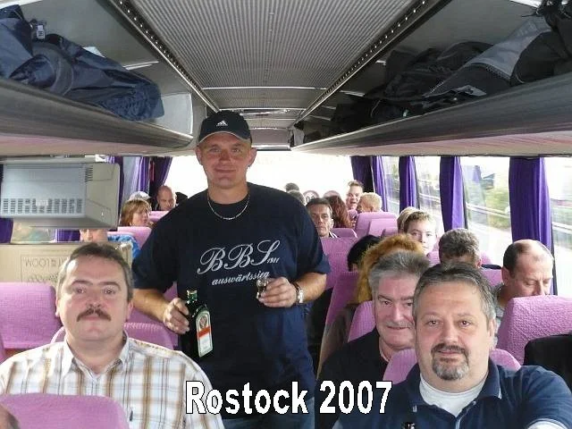 Menschen sitzen in einem Reisebus mit pinken Sitzen. Ein Mann steht in der Mitte und hält zwei Flaschen, alle scheinen eine Gruppenreise zu machen. Es ist im Jahr 2007 in Rostock.