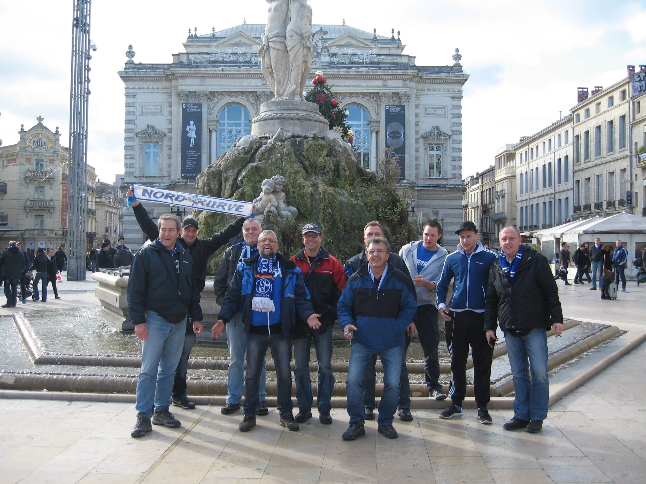 Gruppe von Menschen vor einem Brunnen in einer Stadt, mit einem historischen Gebäude im Hintergrund, trägt Schal mit der Aufschrift 'Nordkurve'.