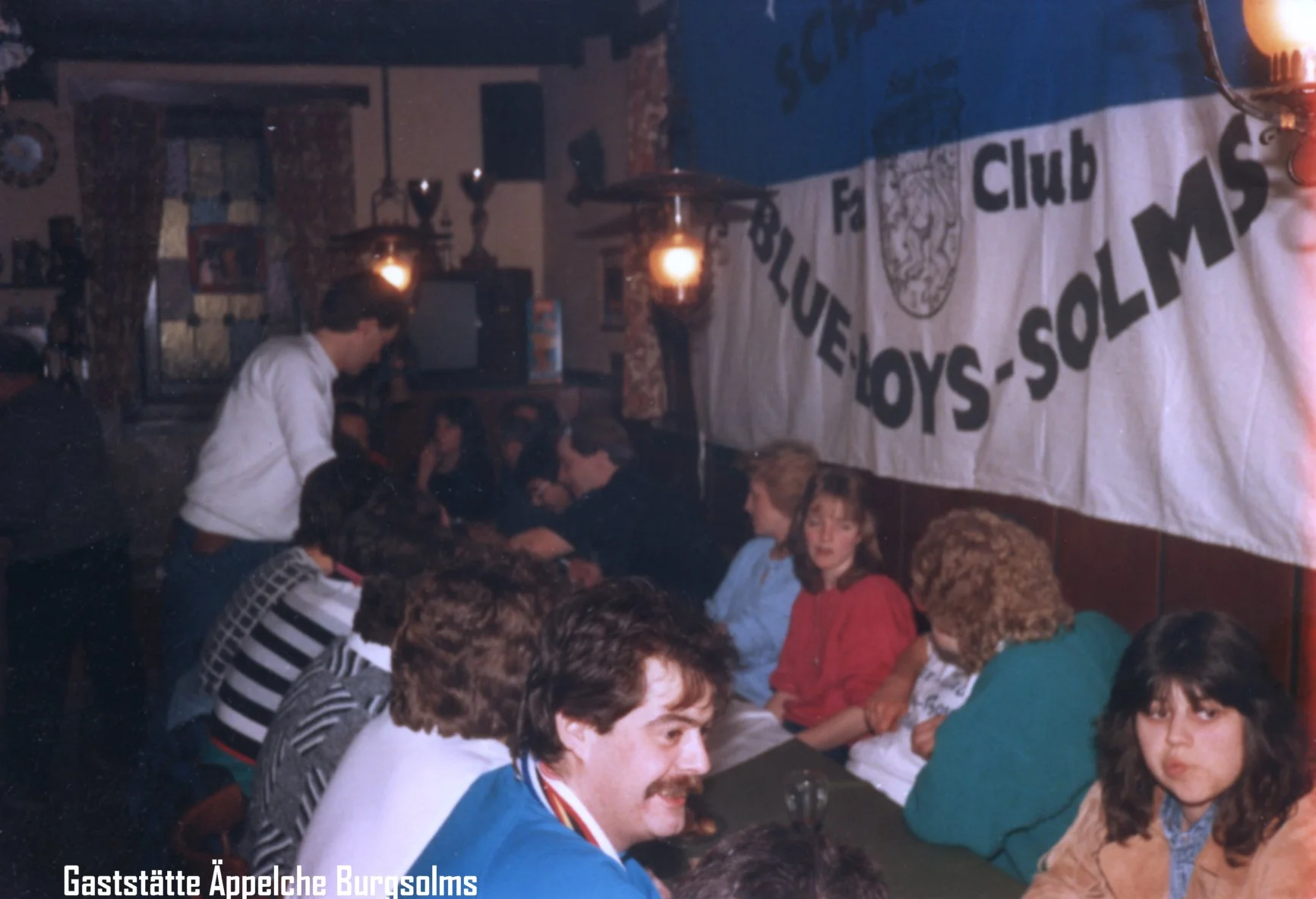 Historische Aufnahme eines Vereins im Lokal Göppingen, Deutschland, mit Menschen, die um Tische sitzen und einen Banner an der Wand, der den Verein 'Fußball-Club Solms' zeigt.