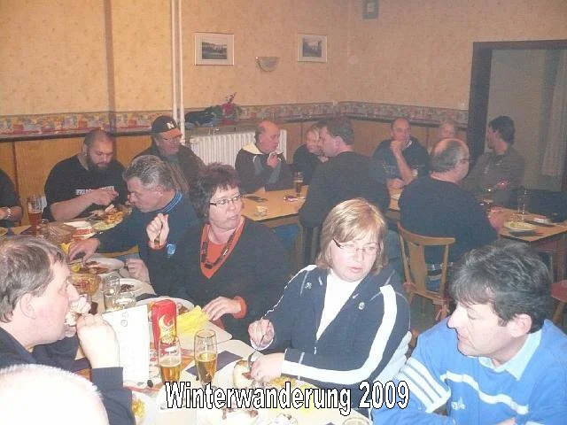 Menschen sitzen an Tischen in einem Restaurant oder einer Bar, essen und trinken. Es ist eine gemütliche Atmosphäre. Der Text auf dem Bild lautet "Winterwanderung 2009".