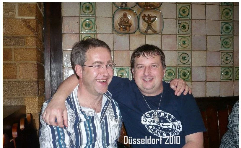 Zwei Männer sitzen in einem Restaurant, lächeln und haben die Arme umeinander gelegt, im Hintergrund Keramikfliesen und Dekorationen an der Wand.