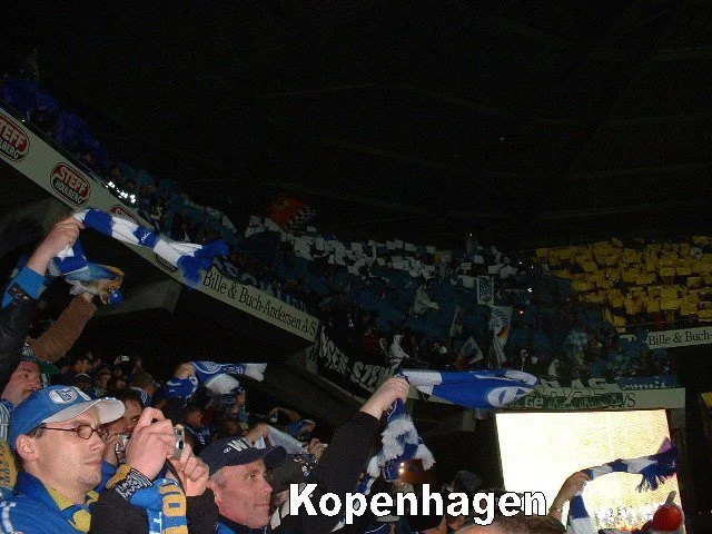 Fans des Kopenhagener Fußballteams beim Spiel im Stadion, einige halten Schal und fotografieren, im Hintergrund sind gelbe und blaue Fahnen und Banner sichtbar.