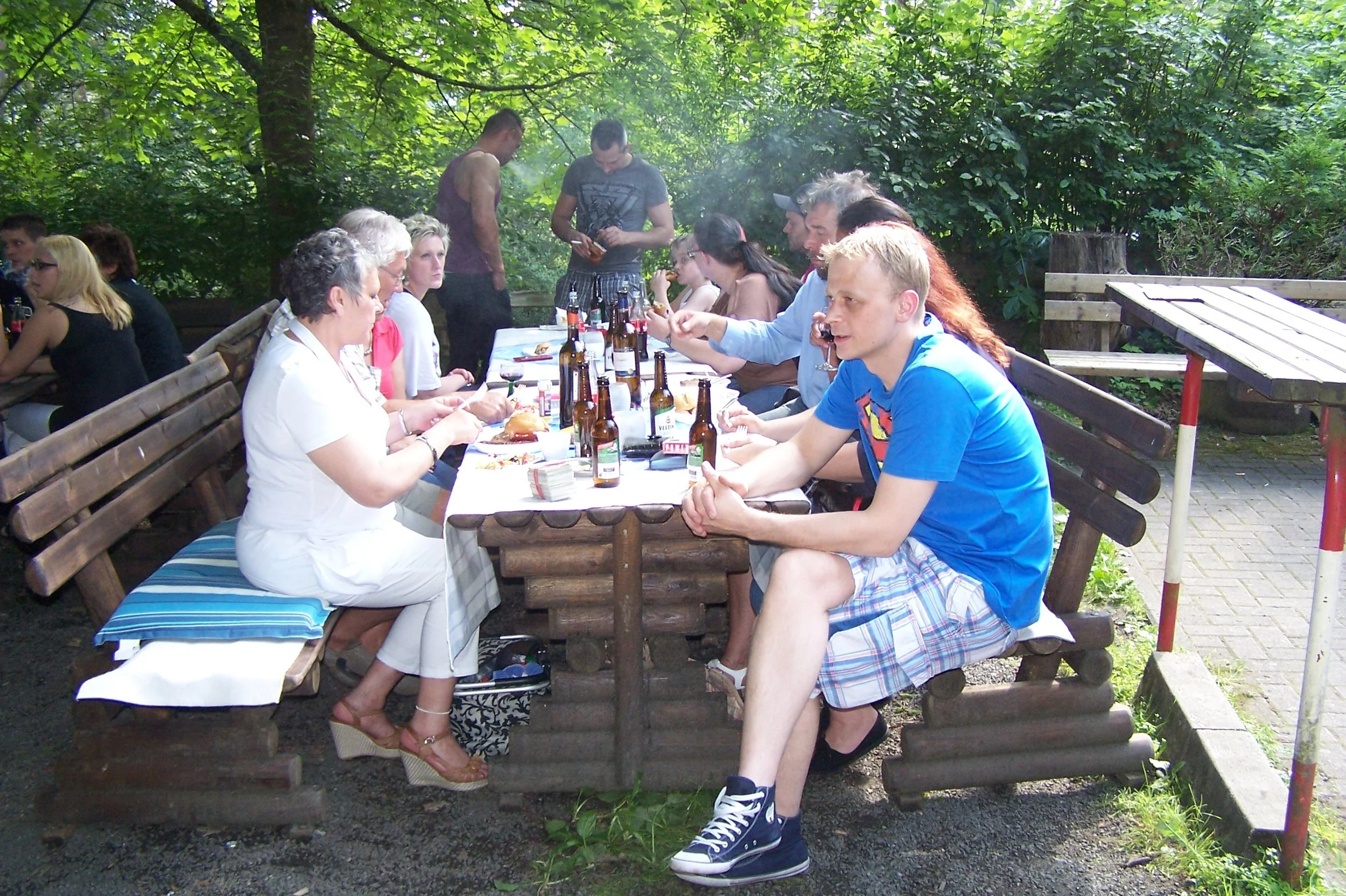 Mehrere Menschen sitzen an einem langen Tisch im Freien, umgeben von Bäumen, während sie essen und trinken.