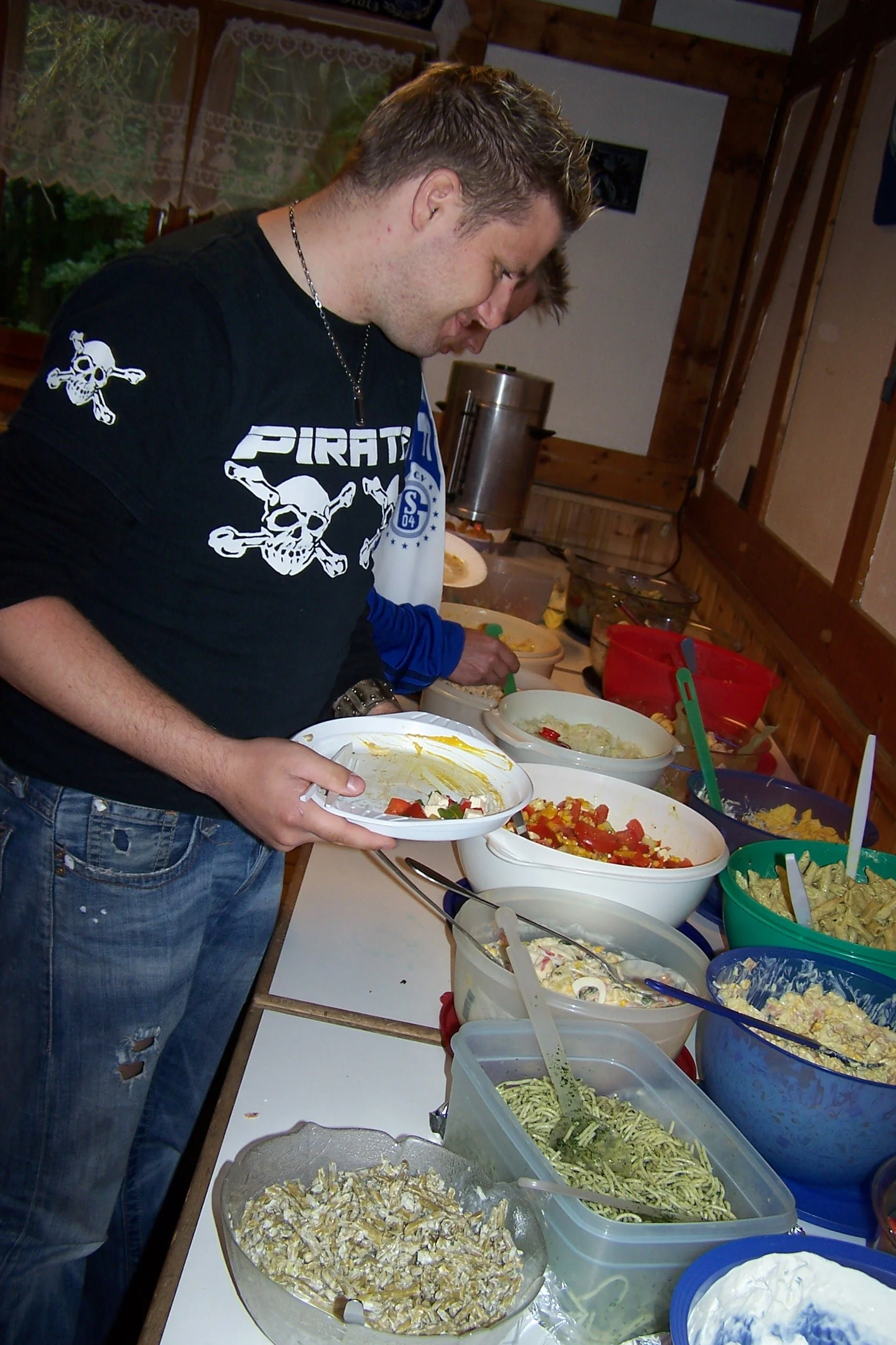 Personen beim Servieren von Salaten und Pasta an einem Buffet in einer rustikalen Holzscheunen-Küche.