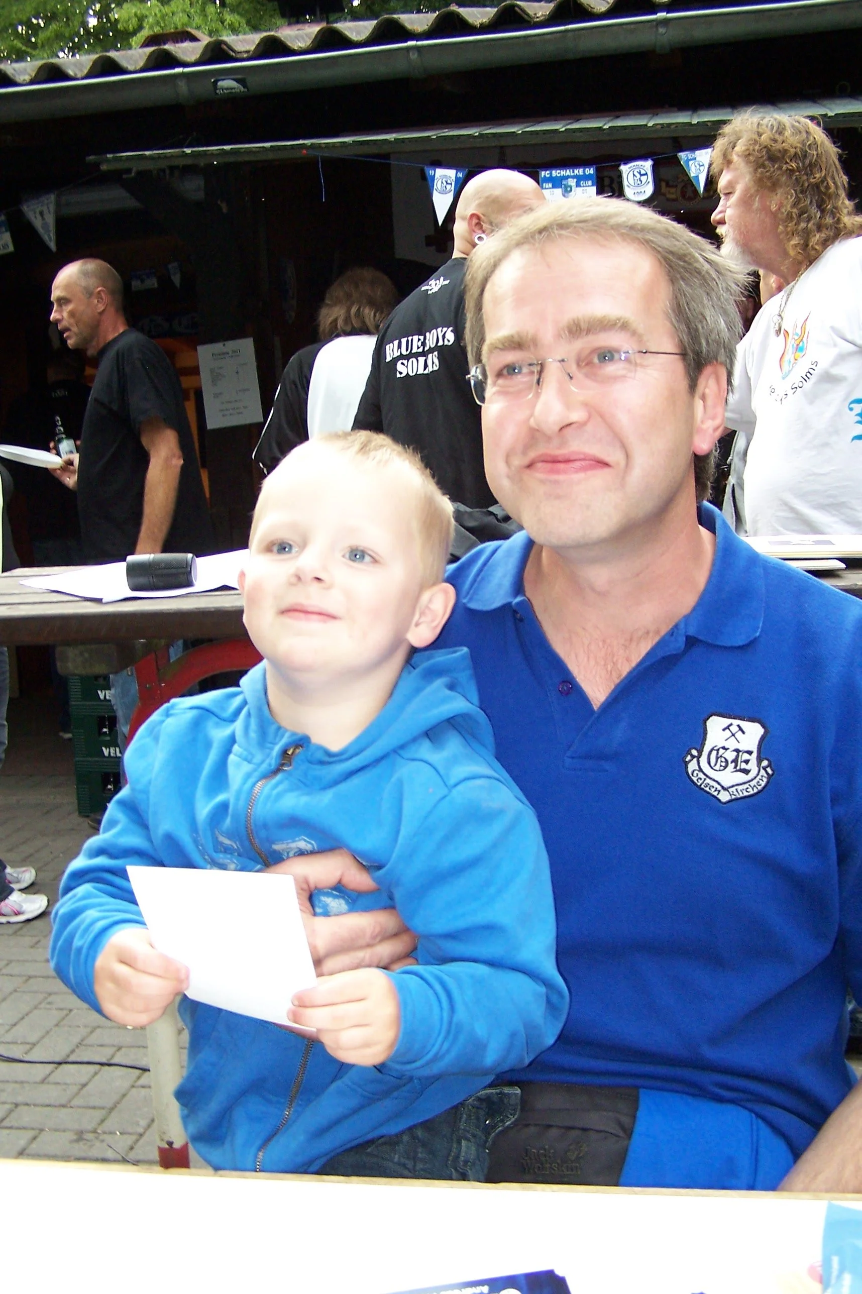 Zwei Männer und ein Kind bei einer Veranstaltung, vielleicht eines Fußballspiels, mit Vereinsamarbeiten. Der Mann auf der rechten Seite trägt ein blauen Polo-Shirt mit einem Vereinslogo, hält das Kind auf dem Schoß, das eine blaue Jacke trägt und ein