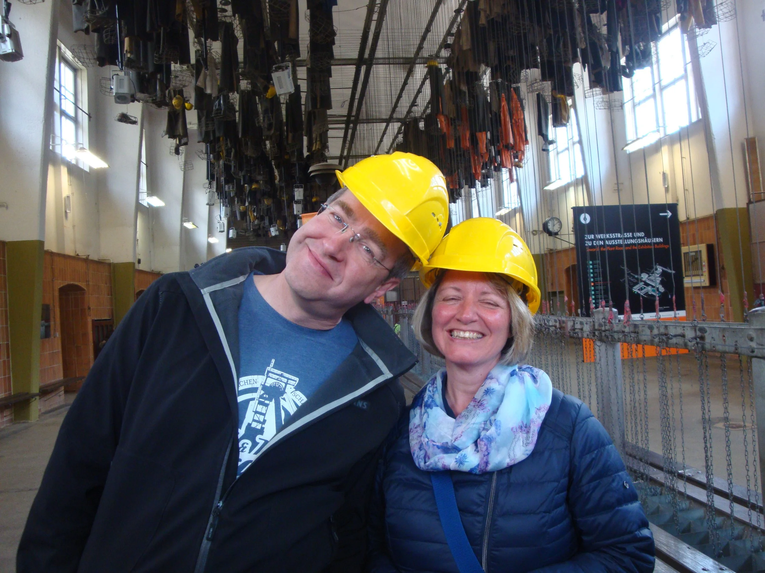 Zwei Menschen tragen Helme in einem industriellen Umfeld mit gestapelten schwarzen und orangefarbenen Materialien im Hintergrund.