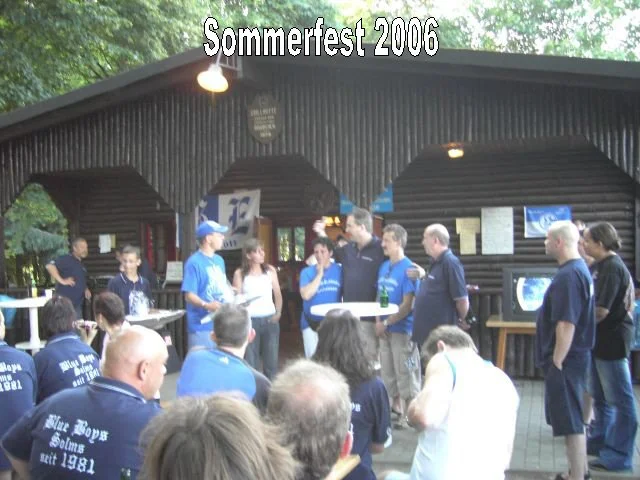 Menschen versammeln sich bei Sommerfest 2006 vor einer Holzhütte, einige sitzen, andere stehen, einige trinken Bier, die Szene findet im Freien statt unter Bäumen.