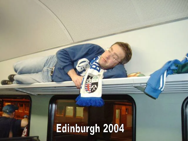 Ein Schlafender Mann in einem Zug, trägt ein blaues Jacke und eine Schal, mit der Aufschrift 'Edinburgh 2004'.