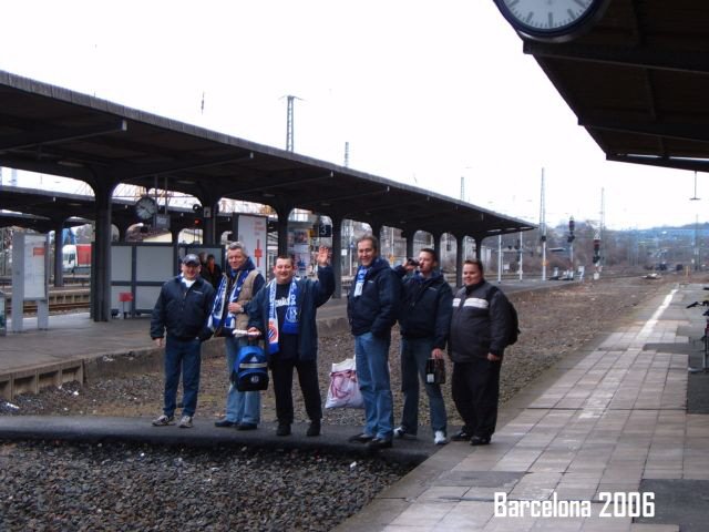 Fünf Menschen stehen auf einem Bahnsteig am Bahnhof in Barcelona im Jahr 2006, einige tragen Jacken und haben Koffer, im Hintergrund sind die Bahngleise und eine Überdachung sichtbar.