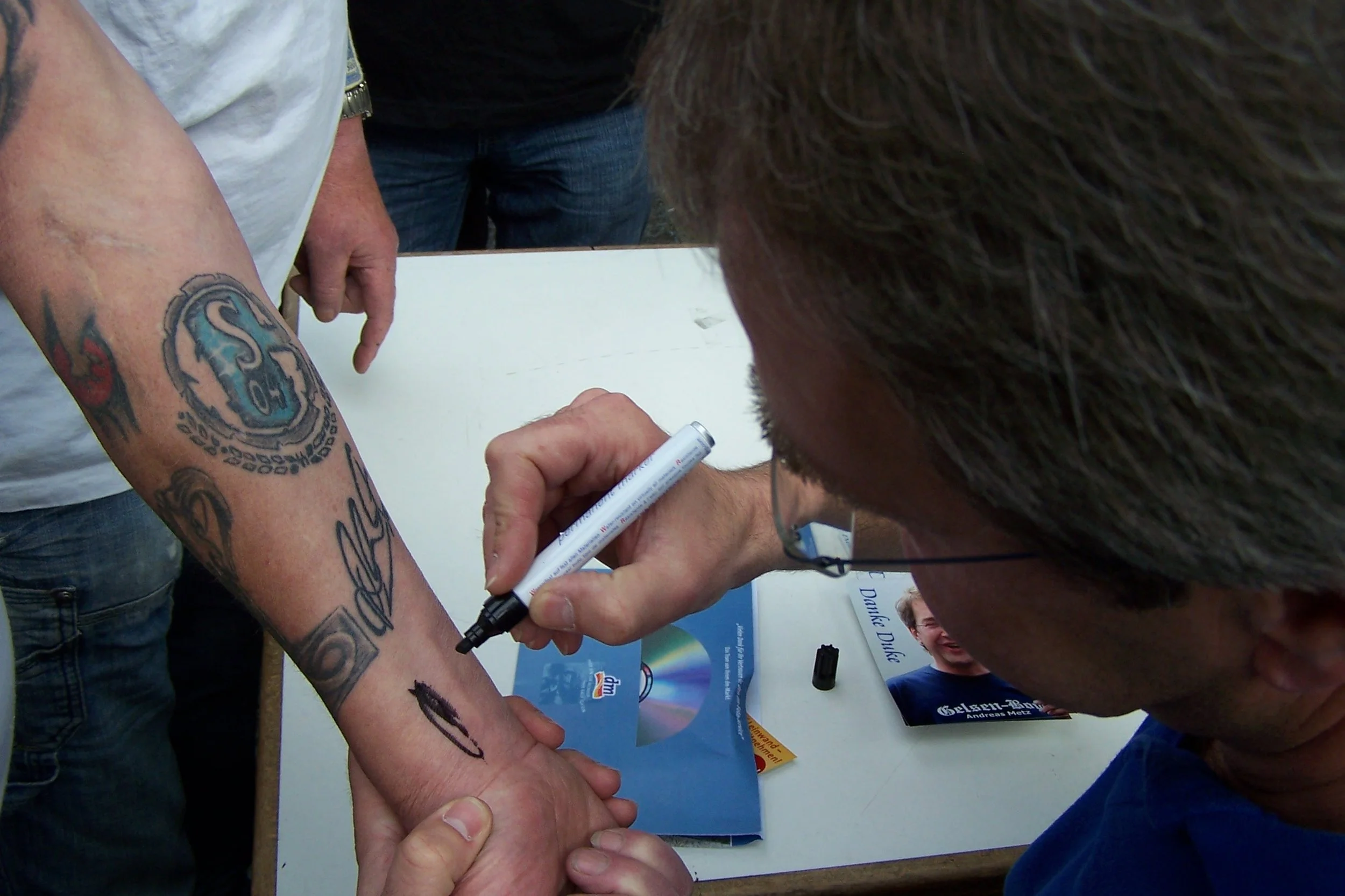 Jemand schreibt mit einem schwarzen Marker auf den Arm einer Person, die Tattoos hat. Es sind mehrere Menschen um einen Tisch herum.