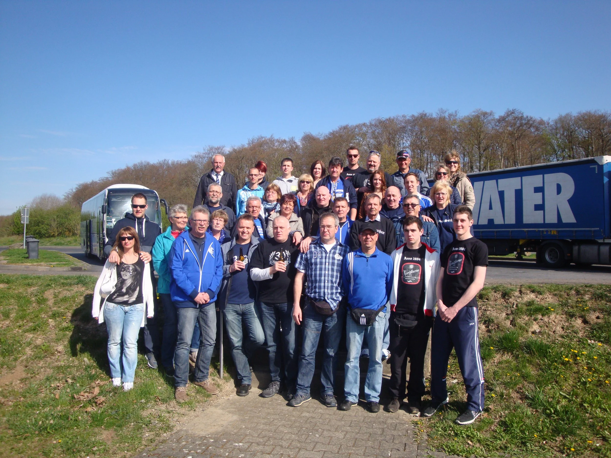 Gruppenfoto von Menschen im Freien vor einem Bus, mit Bäumen im Hintergrund, bei schönem Wetter.