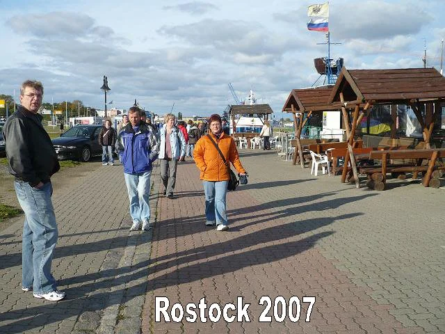 Menschen gehen spazieren an einem Hafen in Rostock im Jahr 2007, mit Holzpavillons und Flaggen, im Hintergrund Schiffe und Kräne.