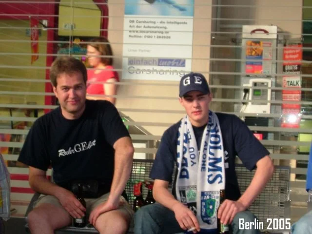 Zwei junge Männer sitzen auf einer Bank in einer öffentlichen Einrichtung, wahrscheinlich 2005 in Berlin, mit einem Hintergrund aus Schaltern und Anzeigen. Beide tragen dunkle T-Shirts, der jüngere Mann trägt eine Mütze und einen Schal.