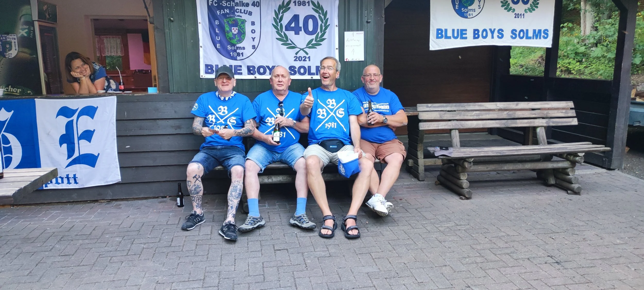 Vier Männer in blauen Trikots sitzen auf einer Bank vor einem Holzgebäude mit Bannern, die den 40. Geburtstag des Blue Boys Solms feiern. Sie trinken Bier, freuen sich und zeigen Gesten der Freude.