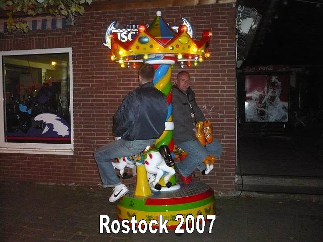Zwei Männer sitzen auf einem Kinderkarussell mit buntem, gestreiftem Mast und Königsfigur oben, vor einem Backstein-Gebäude in Rostock im Jahr 2007.