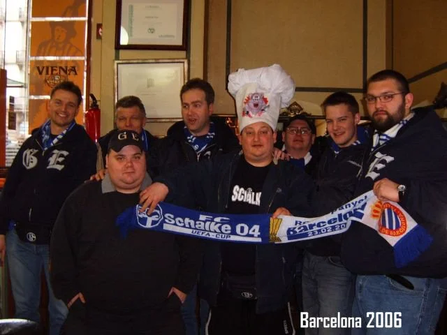 Eine Gruppe von sieben Menschen ist in einem Raum, einer trägt einen Kochhut, alle halten einen Schal der FC Schalke 04, im Hintergrund Poster und Zertifikate. Es ist Barcelona 2006.
