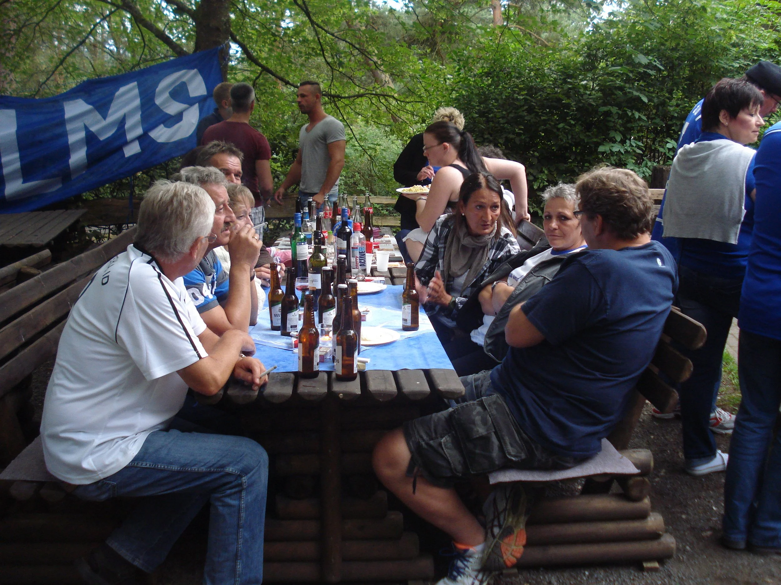 Eine Gruppe von Menschen sitzt an einem langen Holztisch im Freien, umgeben von Bäumen, mit zahlreichen Getränkeflaschen und essen auf dem Tisch, während andere Menschen im Hintergrund stehen und sich unterhalten.