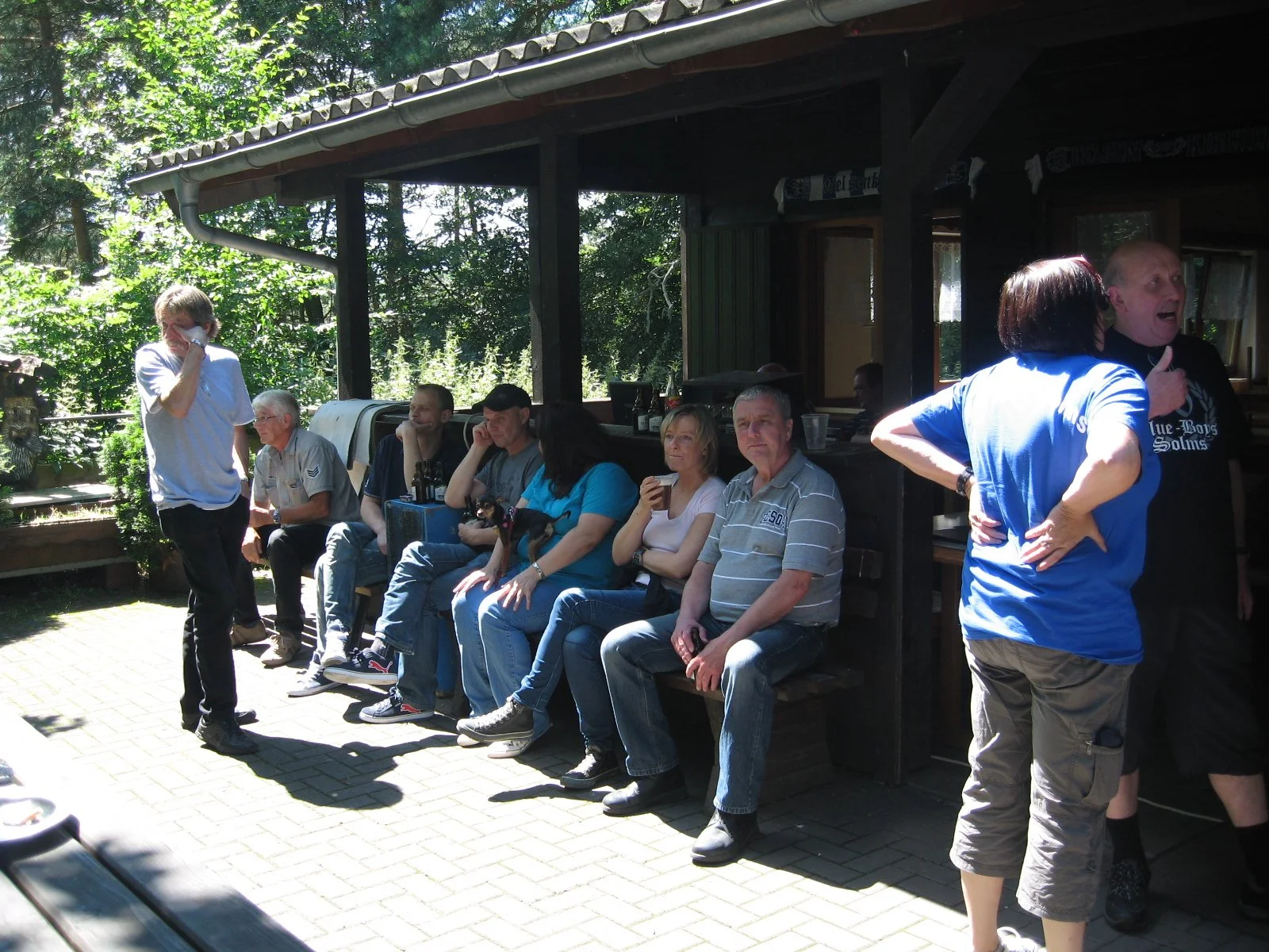 Eine Gruppe von Menschen sitzt und steht vor einem Gebäude im Schatten, einige sind am Abendessen oder Trinken, während andere miteinander sprechen.