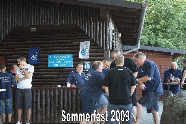 Gruppe von Menschen bei einem Sommerfest 2009, die an einem Holzwagen feiern, einige trinken Kaffee, andere sprechen. Es sind Flags mit FC Schalke 04-Logo im Hintergrund sichtbar.