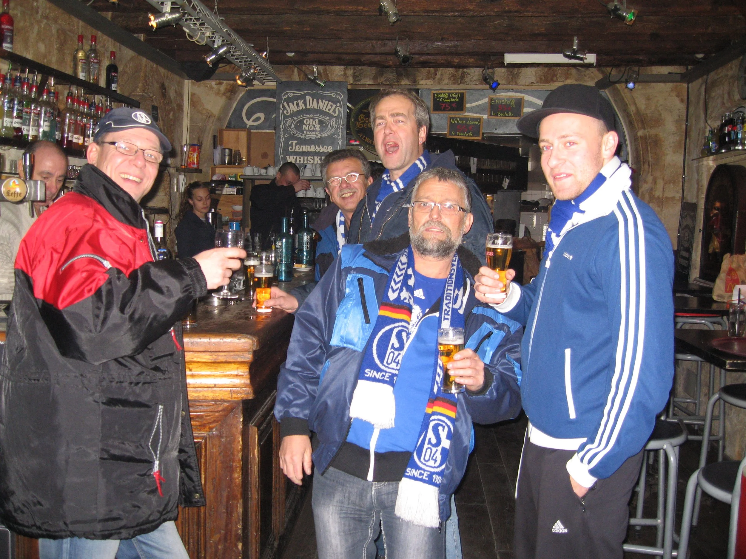 Eine Gruppe von fünf Personen, die in einer Bar oder Kneipe an einem Holztresen stehen, dabei Bier trinken und lächeln. Im Hintergrund sind Regale mit Flaschen und eine Tafel mit Getränkespezialitäten.