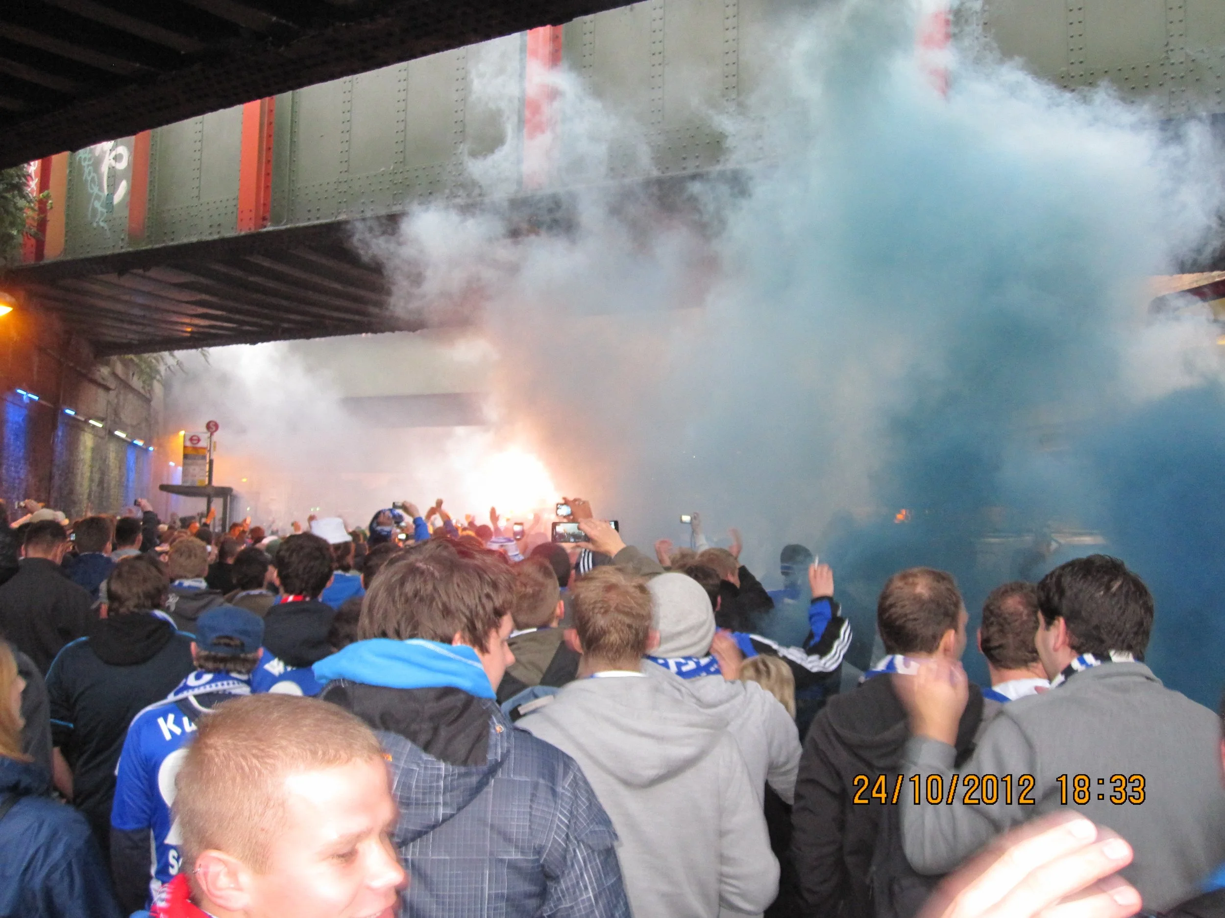 Menschenmenge auf einem Stadion, viele tragen blaue Fußballfansoutfits, hinter ihnen eine Brücke mit Rauch oder Nebel, und die Tageszeit ist abends. Es scheint eine Feier oder ein Spiel zu sein.