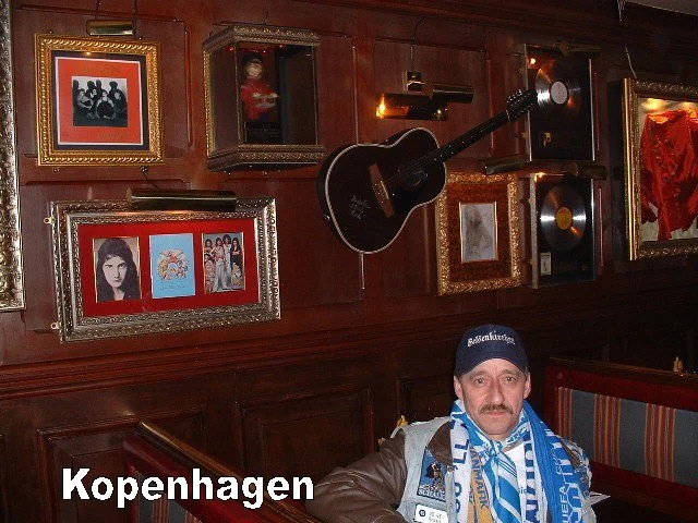 Ein Mann sitzt in einem Pub mit dunkler Holzwand, die mit Bilderrahmen, einem schwarzen Akustik-Gitarren und einem Grammophon dekoriert ist. Der Mann trägt eine blauen Schal und eine Mütze mit dem Wort 'Gelsenkirchen'.