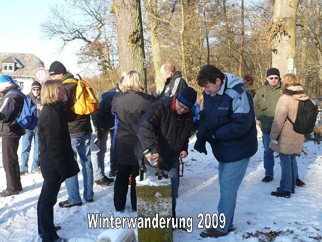 Mehrere Menschen, die Winterkleidung tragen, stehen im Schnee in einem Park mit Bäumen. Zwei Personen in der Mitte schauen auf eine Glasschale, die auf einem Baumstumpf steht. Es scheint eine Winterwanderung im Jahr 2009 zu sein.