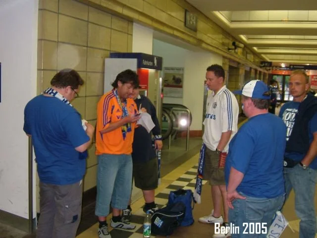 Sechs junge Männer stehen im Flughafen Berlin 2005, einer liest aus einem Papier vor, die anderen hören zu oder schauen. Es sind Trinkflaschen und Taschen auf dem Boden neben ihnen.