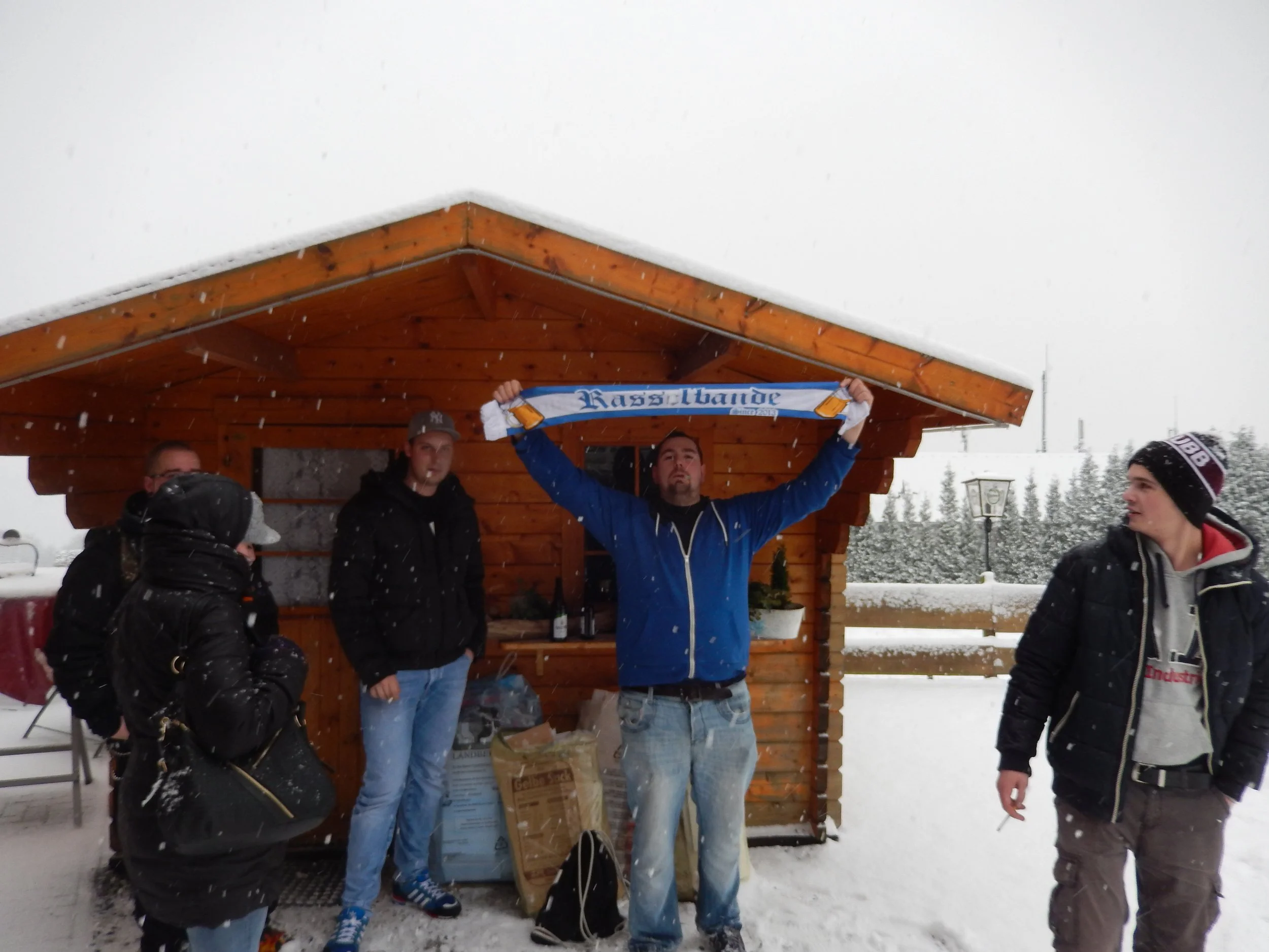 Gruppe von Menschen, die in Schneewetter unter einem Holzdach stehen. Eine Person hält einen Schal mit dem Schriftzug »Kassalator«. Es ist winterlich mit Schnee im Hintergrund.
