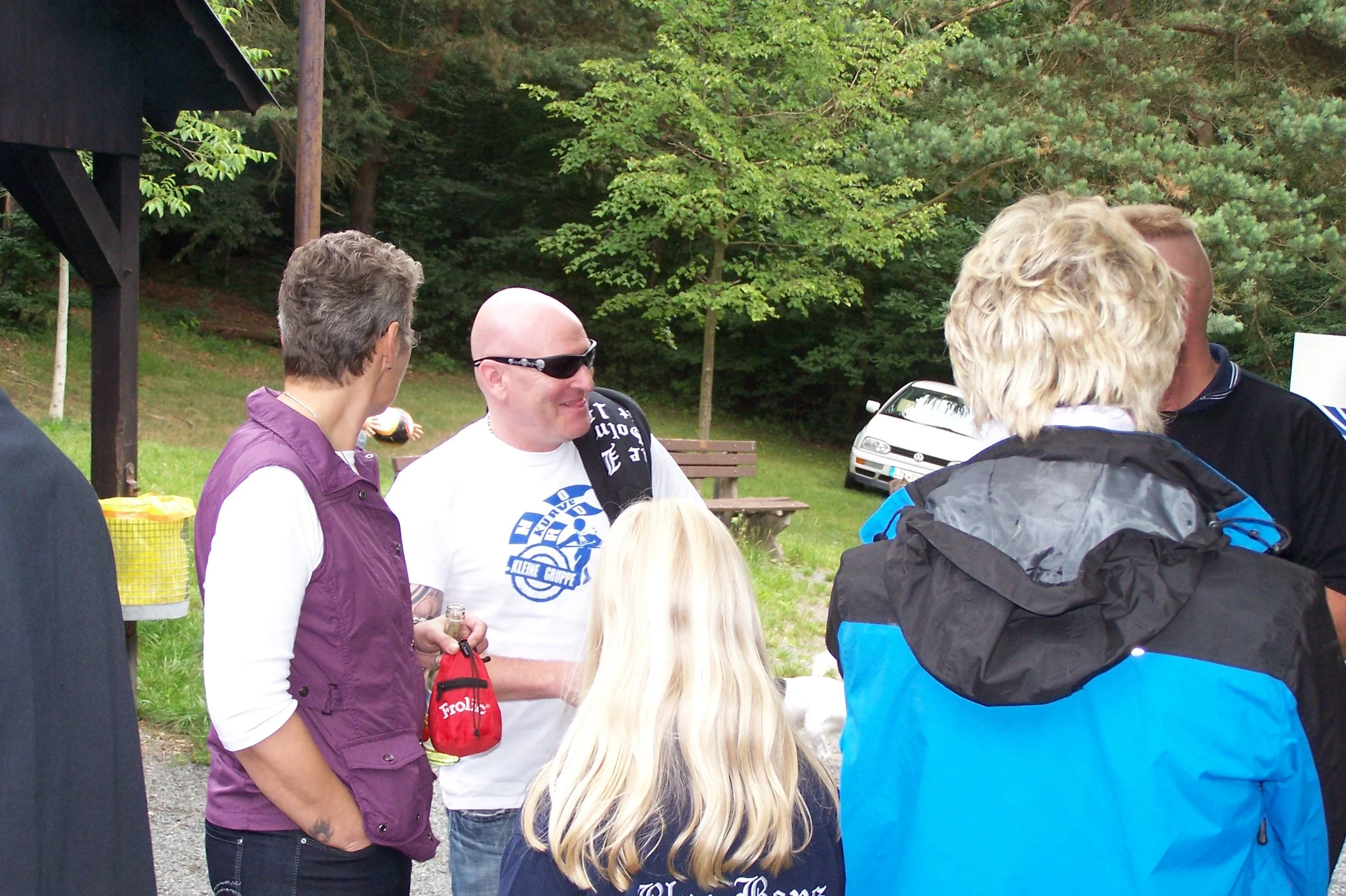 Gruppe von Menschen im Freien, im Hintergrund Bäume und ein Auto, sie scheinen bei einem Treffen oder Event zu sein.
