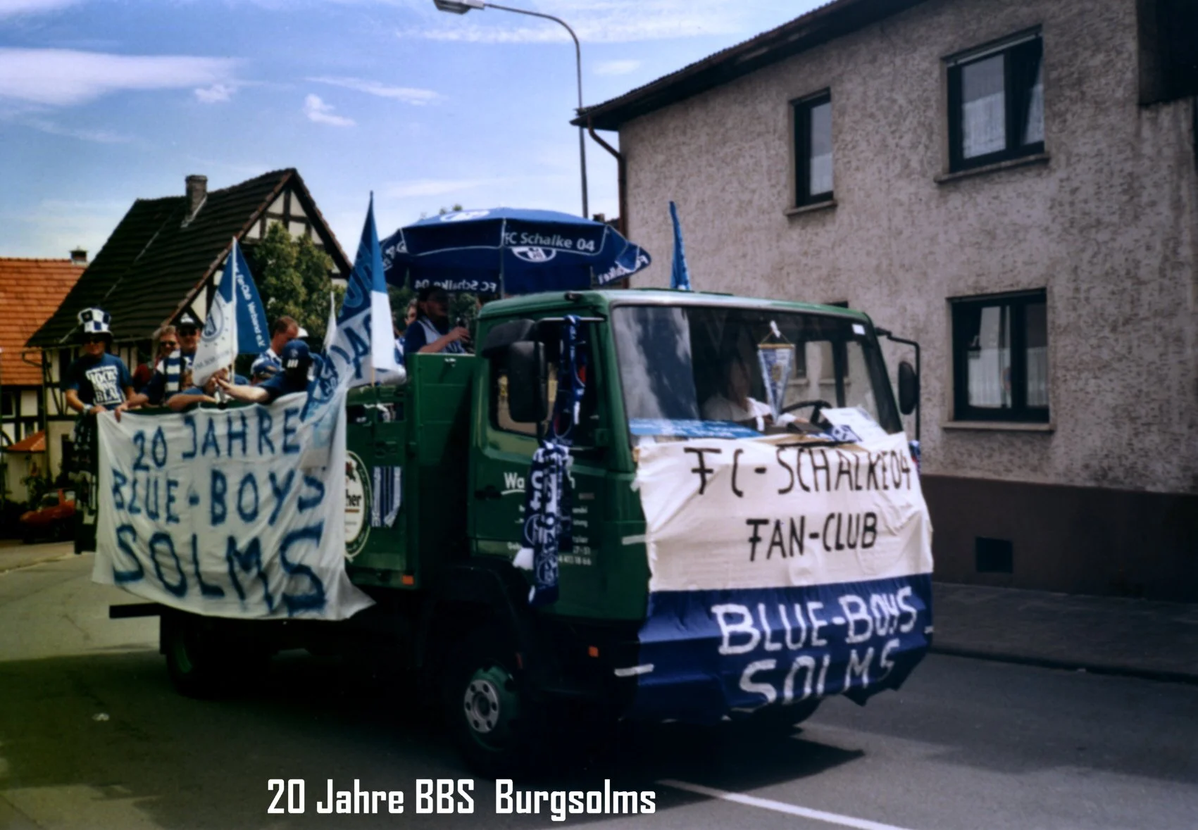 Ein grüner Kleinbus ist mit blauen Fahnen und Bannern geschmückt, auf denen steht '20 Jahre BBS Burgsolms'. Es sind Menschen in blauen Trikots und Mützen, die an einer Parade oder Feier teilnehmen, die die 20-jährige Jubiläumsfeier der BBS Burgsolms 