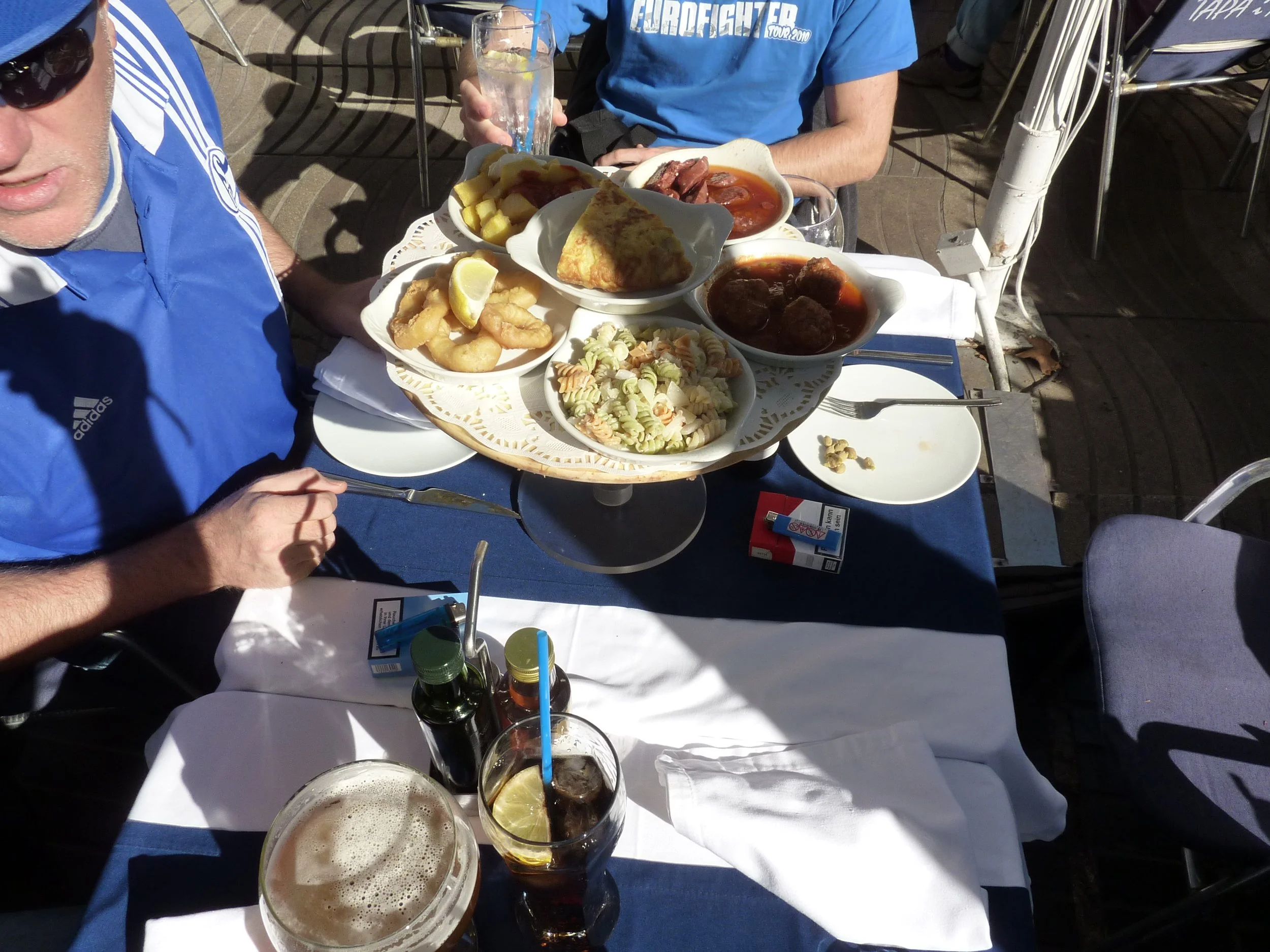 Tisch mit verschiedenen Speisen und Getränken im Freien, Gäste essen und trinken bei Sonnenschein, teilweise sichtbar sind Menschen in blauen Shirts, darunter Pasta, frittiertes Essen, Fleischgerichte und Getränke mit Zitronenscheiben.