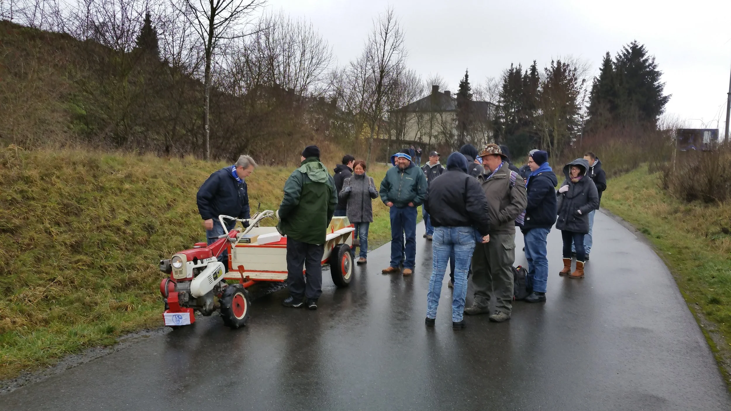 Eine Gruppe Menschen steht auf einer nassen Straße in einer ländlichen Gegend, einige tragen regenkleidung und Hüte, während ein Mann einen kleinen Traktor mit Anbaugerät zeigt.
