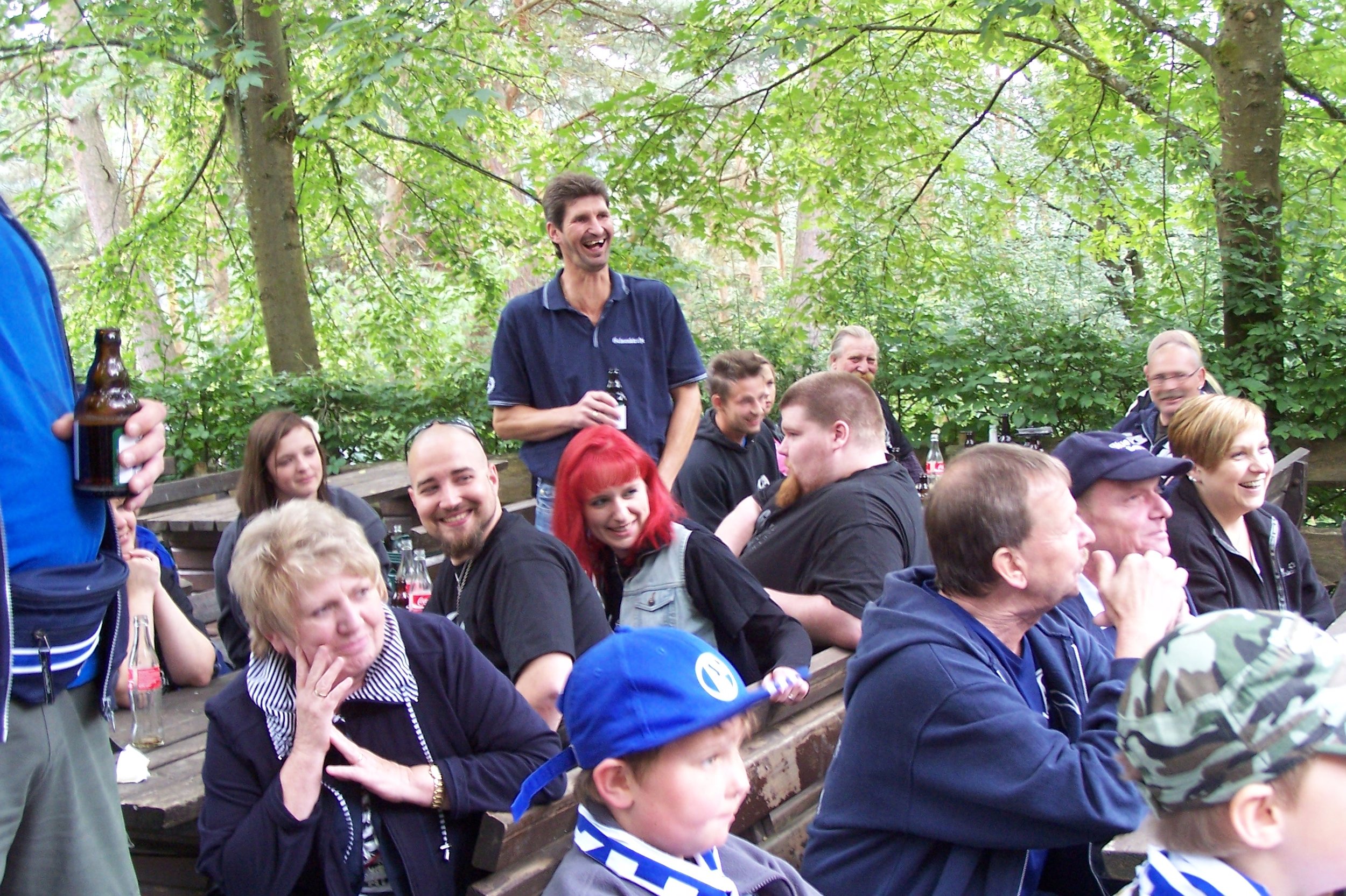 Gruppe fröhlicher Menschen sitzt an einem Tisch im Freien, einige lachen und trinken Bier, im Hintergrund Bäume und grüne Blätter.