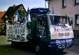 Bunter Umzugswagen mit Bannern für eine Fußballveranstaltung, in einem Wohngebiet, mit Häusern im Hintergrund.