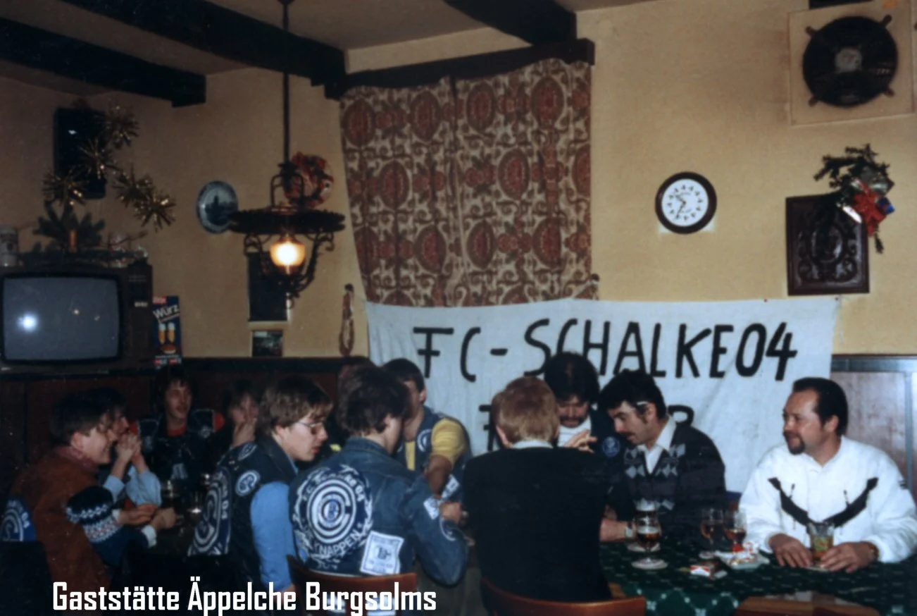 Eine Gruppe von Menschen sitzt an einem Tisch in einer Gaststätte, mit einem Banner im Hintergrund, das 'FC-SCHALKE04' liest. Es gibt einen alten Fernseher, Wanddekoration und Weihnachtsdekoration.