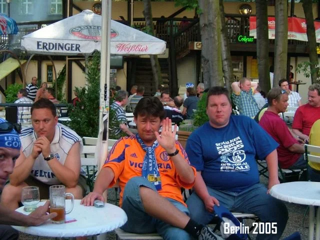 Gruppe von Menschen, die draußen in einer Bar oder einem Biergarten sitzen. Es gibt Tische, Gläser und einen großen Sonnenschirm mit dem Aufdruck "Erdinger". Im Hintergrund sind weitere Gäste und ein mehrstöckiges Gebäude. Der Text "Berlin 2005" ist 