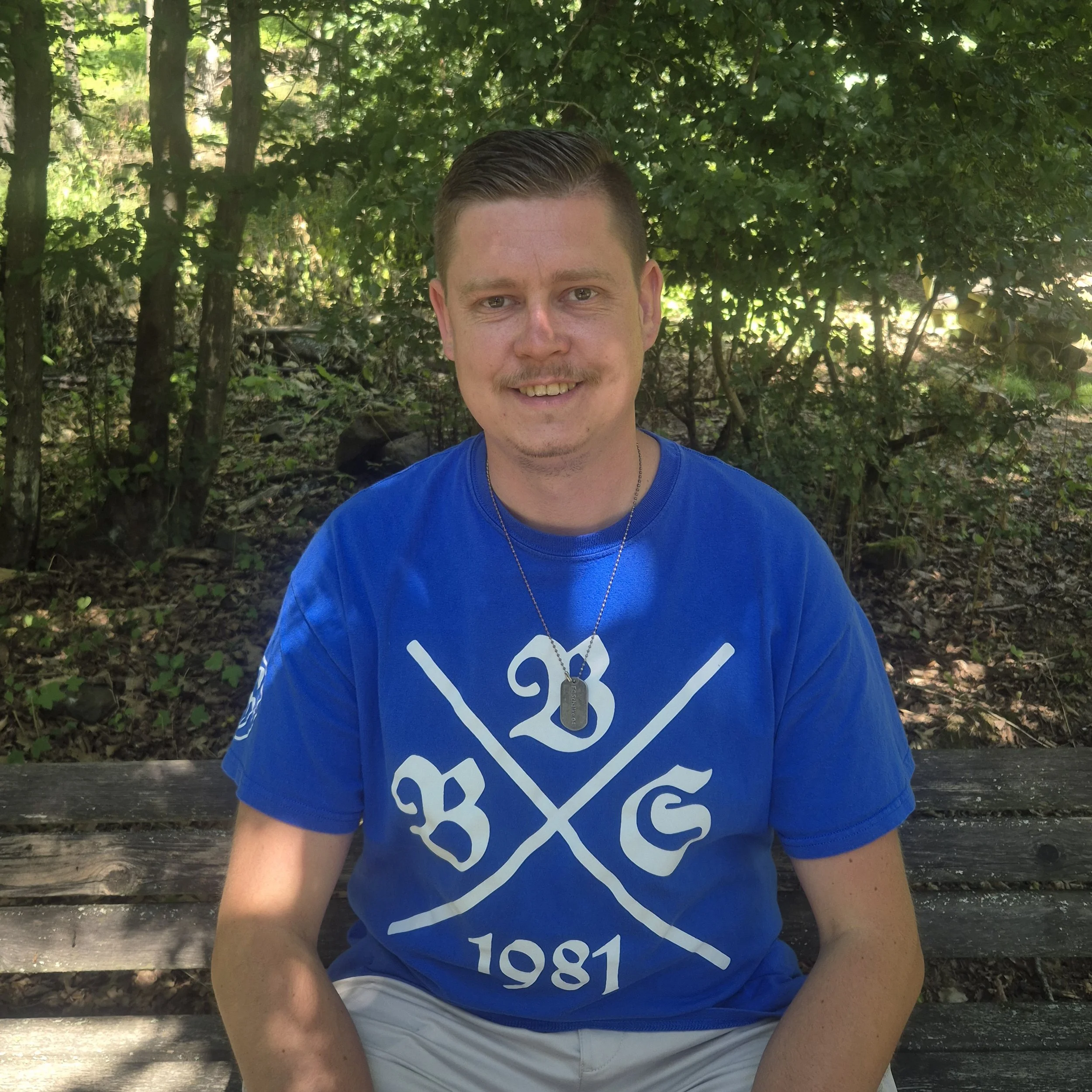 Ein Mann mit kurzgeschnittenen Haaren und einem freundlichen Lächeln, getragen in einem blauen T-Shirt mit weißen Druck, sitzt auf einer einfachen Bank im Freien, umgeben von dichtem grünen Laub und Bäumen.