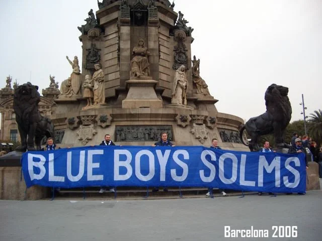 Menschen halten ein blaues Banner mit der Aufschrift 'BLUE BOYS SOLMS' vor einer monumentalen Skulptur in Barcelona, 2006.
