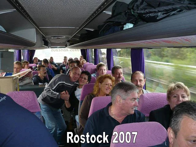 Menschen sitzen in einem Reisebus mit pinken Sitzen. Einige Menschen sitzen, andere stehen. Es gibt Lichterketten und Vorhänge im Fenster. Der Bus ist voll. Auf dem Bild ist eine Gruppe von Personen, die lachen und posieren. Der Blick nach vorne ist 