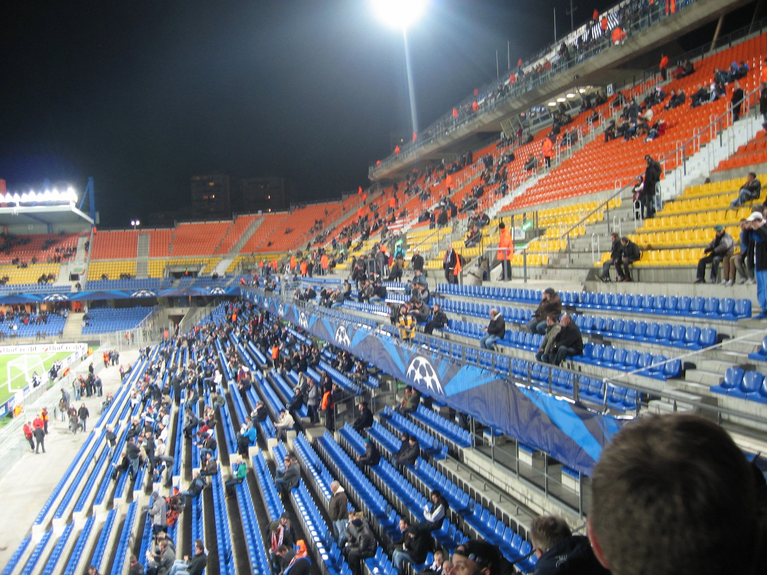 Blick in ein leeres Stadion bei Nacht mit wenigen Zuschauern auf den orangefarbenen und blauen Sitzplätzen, einige Personen sitzen und stehen verteilt