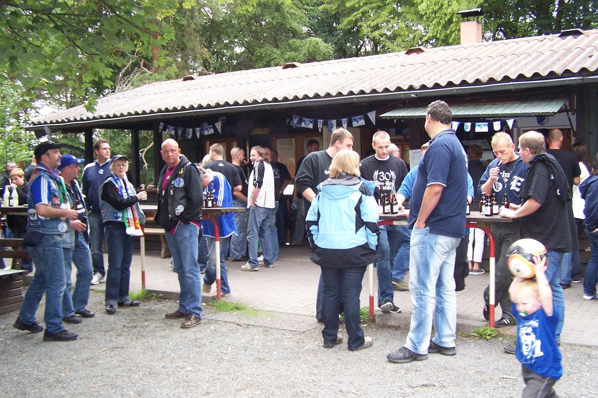 Gruppe von Menschen im Freien vor einem Holzgebäude, die Getränke trinken und sich unterhalten, mit einigen im Fußballtrikot, umgeben von Bäumen.