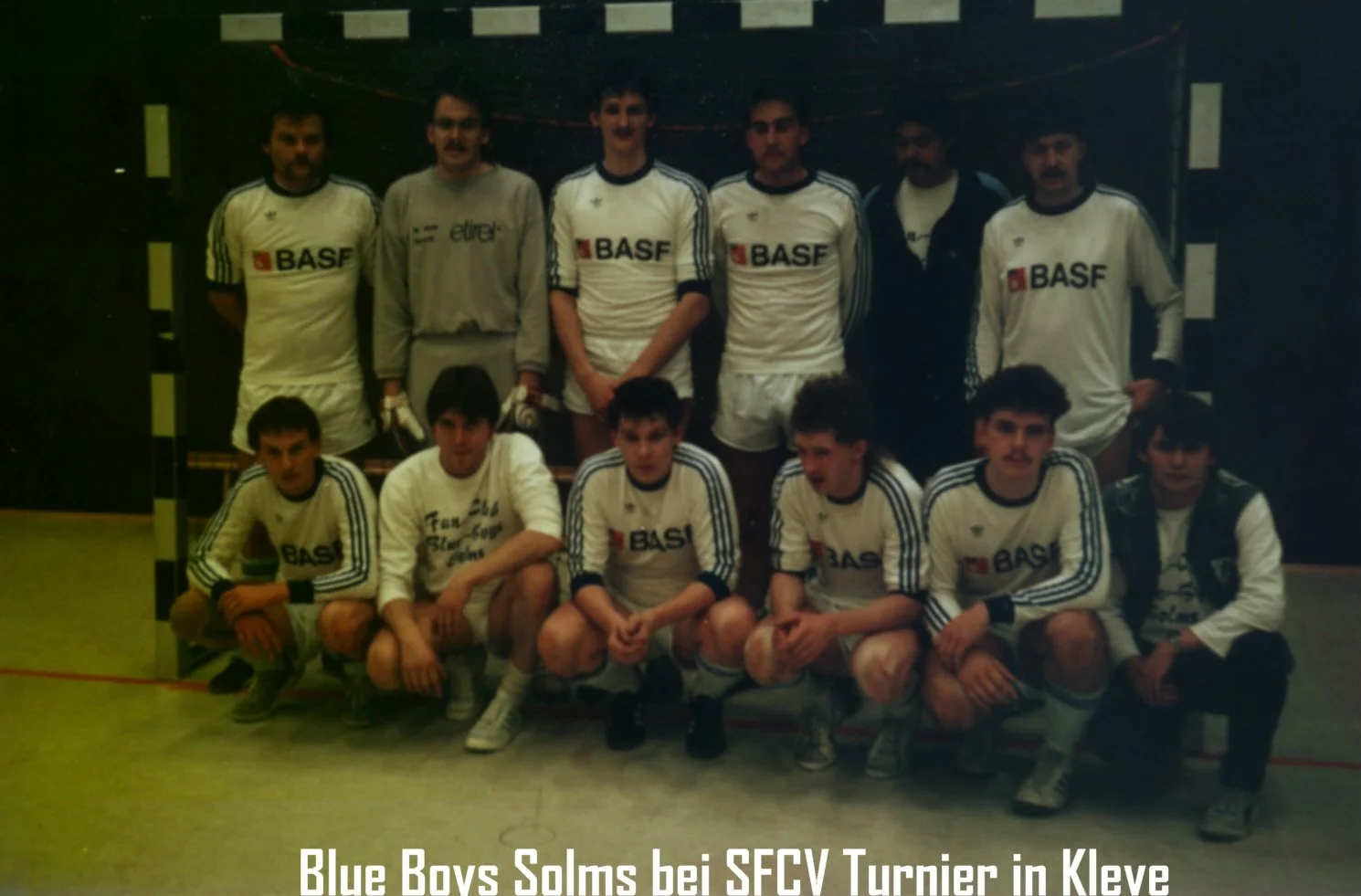 Gruppenfoto von einem Jugendfußballteam namens Blue Boys Solms beim SFCV Turnier in Kleve, alle Spieler tragen weiße Trikots mit BASF-Logo, einige stehen im Hintergrund, einige sitzen im Vordergrund.